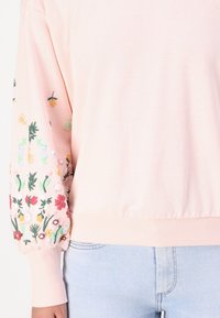 Felpa rosa chiaro con ricami floreali sulle maniche, polsini e orlo a coste. Tessuto morbido, vestibilità leggermente rilassata, con motivi floreali colorati.