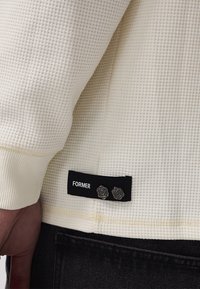White textured sweatshirt with a waffle knit pattern, featuring a black label that reads "FORMER" and decorative silver rose buttons.