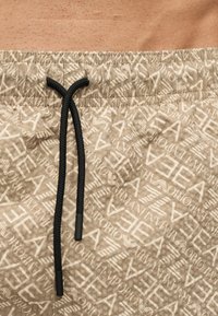 Nærbilde av beige badeshorts med hvite EA7 EMPORIO ARMANI-logoer over hele og en svart snor i den elastiske linningen.
