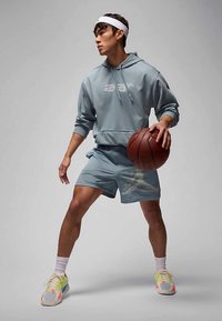 Camisola com capuz azul-claro com gráfico frontal, calções azuis a condizer com padrão texturizado e sapatilhas multicoloridas. Segurando uma bola de basquete.