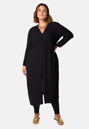 Zwarte lange cardigan met V-hals, lange mouwen en een voorkant met split. Draagt over zwarte leggings, gecombineerd met gouden slippers. Textuurstof.