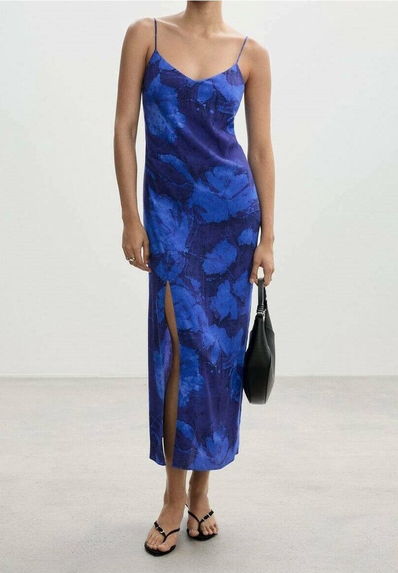 Femme portant une robe midi bleue à fleurs avec une fente haute sur la cuisse, tenant un sac à main noir et portant des sandales noires.