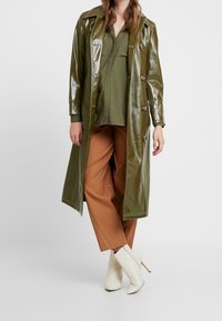 Cappotto lungo verde oliva lucido con bottoni doppi, abbinato a una camicia verde oliva larga e pantaloni marroni; stivaletti bianchi a punta completano il look.