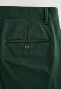 Pantalon en tissu vert foncé avec une poche passepoilée à l'arrière, fermée par un seul bouton sombre et une boucle de ceinture à la taille.
