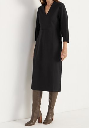 Frau in einem schwarzen V-Ausschnitt, langärmligen Midikleid, kombiniert mit langen, taupefarbenen Lederstiefeln, steht vor einem weißen Hintergrund.