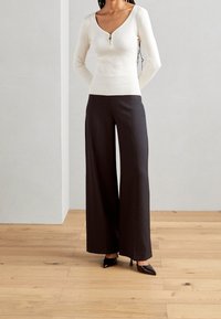 Maglione a coste crema con maniche lunghe e colletto con zip, abbinato a pantaloni neri a gamba larga e tacchi a punta neri. Sfondo in legno.