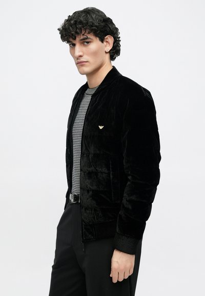 Μαύρο βελούδινο μπουφάν τύπου bomber με ριμπ μανσέτες και τελείωμα, με χρυσή λεπτομέρεια λογότυπου στο στήθος, φορεμένο πάνω από μια ριγέ πουκάμισο.