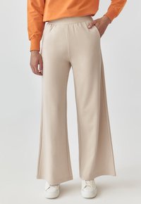 Pantaloni bej cu croi larg, cu talie elastică, textură netedă, buzunare laterale și o potrivire lejeră. Asortați cu adidași albi.