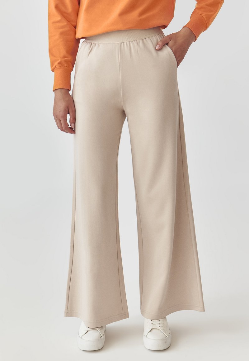 Pantaloni bej cu croi larg, cu talie elastică, textură netedă, buzunare laterale și o potrivire lejeră. Asortați cu adidași albi.