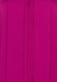 Blouse fuchsia avec des plis verticaux, dotée d'une rangée de boutons ronds au centre. Texture de tissu lisse avec des détails de couture subtils.