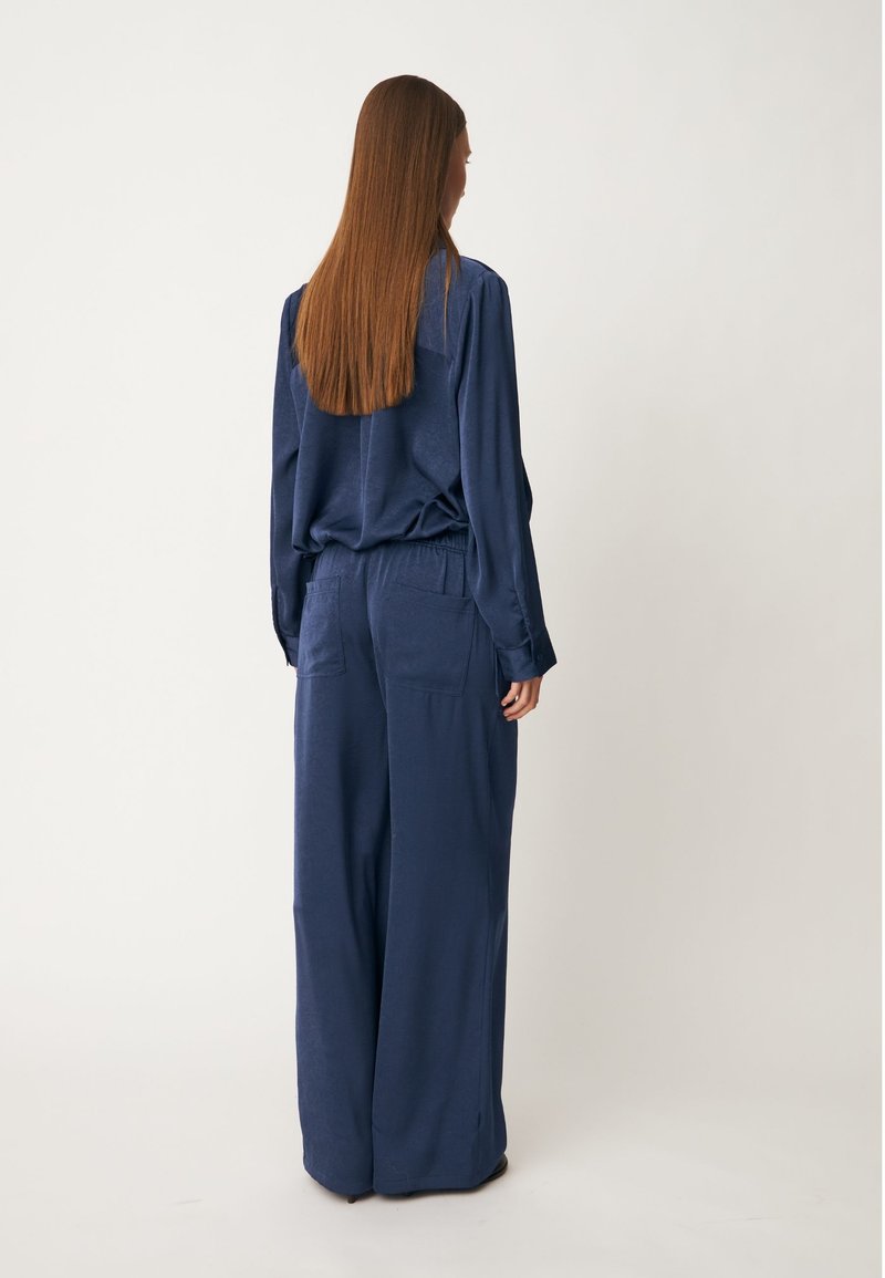 Personne aux longs cheveux bruns portant une chemise ample bleu marine à manches longues et un pantalon à jambes larges, debout de dos sur un fond uni.