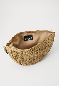 Tkaná slaměná taška v béžové barvě s texturovaným povrchem a hladkým, hnědým látkovým podšívkou. Obsahuje černou nálepku "PIECES" a zlatý zip.