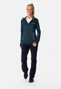 Giacca a zip blu scuro con cappuccio, caratterizzata da dettagli bianchi e tasche laterali, indossata con pantaloni scuri e scarpe da trekking. Design semplice e atletico.