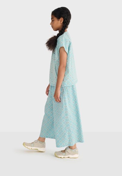 Fille aux cheveux tressés marchant, portant un haut à manches courtes à motifs bleus et un pantalon large assorti, avec des baskets beiges.