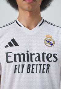 La maglia da calcio bianca presenta un design a scollo a V, dettagli neri e un motivo testurizzato. Mostra "Emirates FLY BETTER" e il logo del Real Madrid.