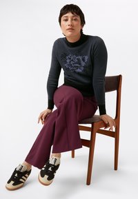 Maglione lavorato a maglia blu navy con motivo a pois e design floreale, abbinato a pantaloni svasati bordeaux e sneakers nere e beige. Seduta su una sedia di legno.