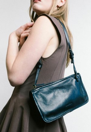 Borsa a tracolla in pelle nera con texture liscia, una tracolla annodata e forma rettangolare, indossata sulla spalla, su uno sfondo neutro.