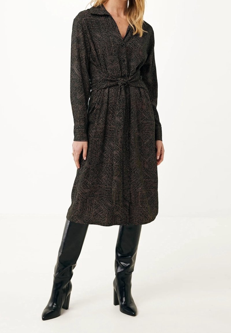 Femme portant une robe longue à manches longues à motifs avec ceinture, associée à des bottes en cuir noires montant jusqu'aux genoux, debout devant un fond uni.