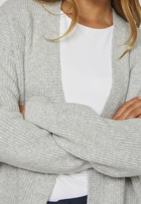 Světlě šedý pletený kardigan s žebrovanou strukturou, otevřený vpředu a s dlouhými rukávy, vrstvený přes bílý top. Měkký materiál, ležérní design.
