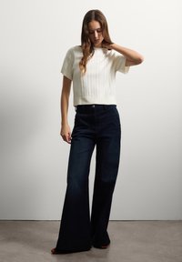 Witte geribbelde cropped trui gecombineerd met donkerblauwe wijdvallende jeans, met knoopsluiting en subtiele textuur. Model staat tegen een effen achtergrond.