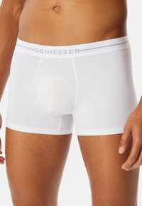 Mandligt torso iført hvide boxer briefs fra Schiesser, som viser taljebånd og øvre lår mod en ensfarvet baggrund.