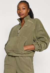 Sudadera de forro polar verde oliva con media cremallera, cuello alto, gran bolsillo frontal y puños elásticos. Presenta un sutil detalle de costuras y un logotipo.