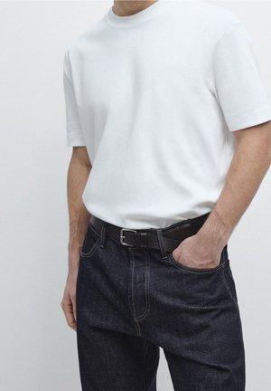 Homme portant un t-shirt blanc uni à manches courtes, un jean bleu foncé et une ceinture noire, une main dans la poche, faisant face à l’avant.
