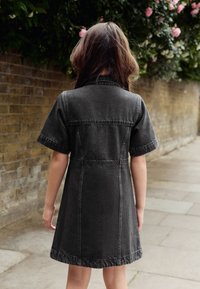 Robe en denim noir à manches courtes, coutures structurées et jupe légèrement évasée, vue de dos devant un mur de briques.