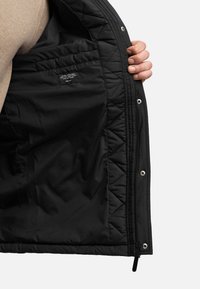 Zwarte gequiltte jas binnenzijde met zichtbare stiksels en een label. Bevat een rits en drukknoopjes langs de opening aan de voorkant.