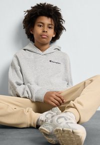 Gray hoodie with a logo on the chest, paired with beige sweatpants. The individual is seated with a neutral expression and textured curly hair.