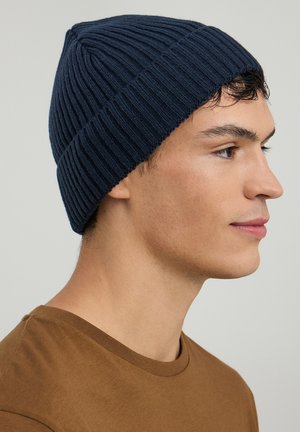 Beanie a coste blu navy con orlo arrotolato, caratterizzato da un design testurizzato. Indossato da una persona con una camicia marrone neutra.