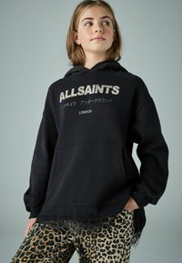 Čierna mikina s kapucňou a grafickým logom v metalickom písmene, skombinovaná so zelenými nohavicami s leopardím vzorom a čipkovým lemom na okraji.