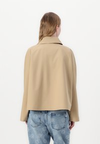Giacca beige corta con colletto ampio e maniche lunghe, realizzata in tessuto morbido. Indossata sopra jeans blu a fantasia, vista da dietro.