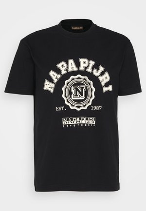 Person iført en sort Napapijri-logo t-shirt, blå jeans og hvide sneakers, der står mod en ensfarvet hvid baggrund.