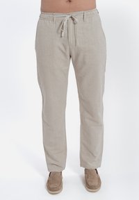 Pantaloni in lino beige con vita elasticizzata e coulisse, dotati di tasche laterali e vestibilità rilassata, abbinati a scarpe marroni senza lacci.