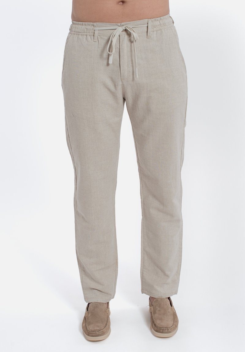Pantaloni in lino beige con vita elasticizzata e coulisse, dotati di tasche laterali e vestibilità rilassata, abbinati a scarpe marroni senza lacci.