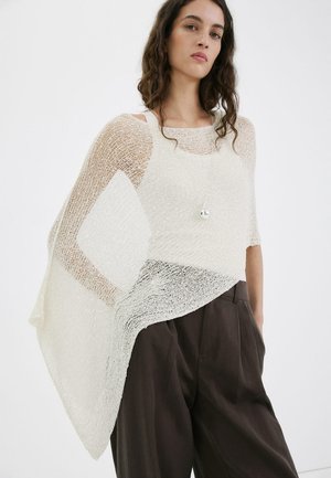 Mladá žena v voľnom, priesvitnom krémovom ponču cez biely top a tmavé široké nohavice na jednoduchom pozadí.