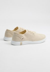 Zapatillas de lona beige con suela de goma blanca, punta redonda y tres ojetes. La parte superior texturizada presenta salpicaduras sutiles; logo de acento marrón en el lateral.