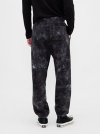 Άνδρας φορώντας μαύρα και γκρίζα tie-dye παντελόνια jogger με πίσω τσέπη, συνδυασμένα με μαύρα αθλητικά παπούτσια με λευκές σόλες, στέκεται σε άσπρο φόντο.