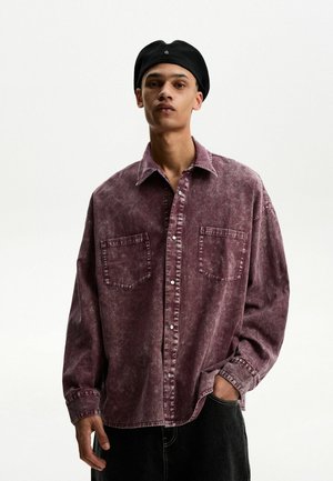 Langarm, ausgewaschenes bordeauxrotes Denim-Hemd mit Knopfverschluss, zwei Brusttaschen und lockerer Passform. Über schwarzer Hose getragen.