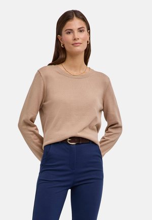 Vrouw met lang bruin haar, die een beige trui, gouden oorbellen, een ketting en donkerblauwe broek met een bruine riem draagt, staand tegen een effen achtergrond.