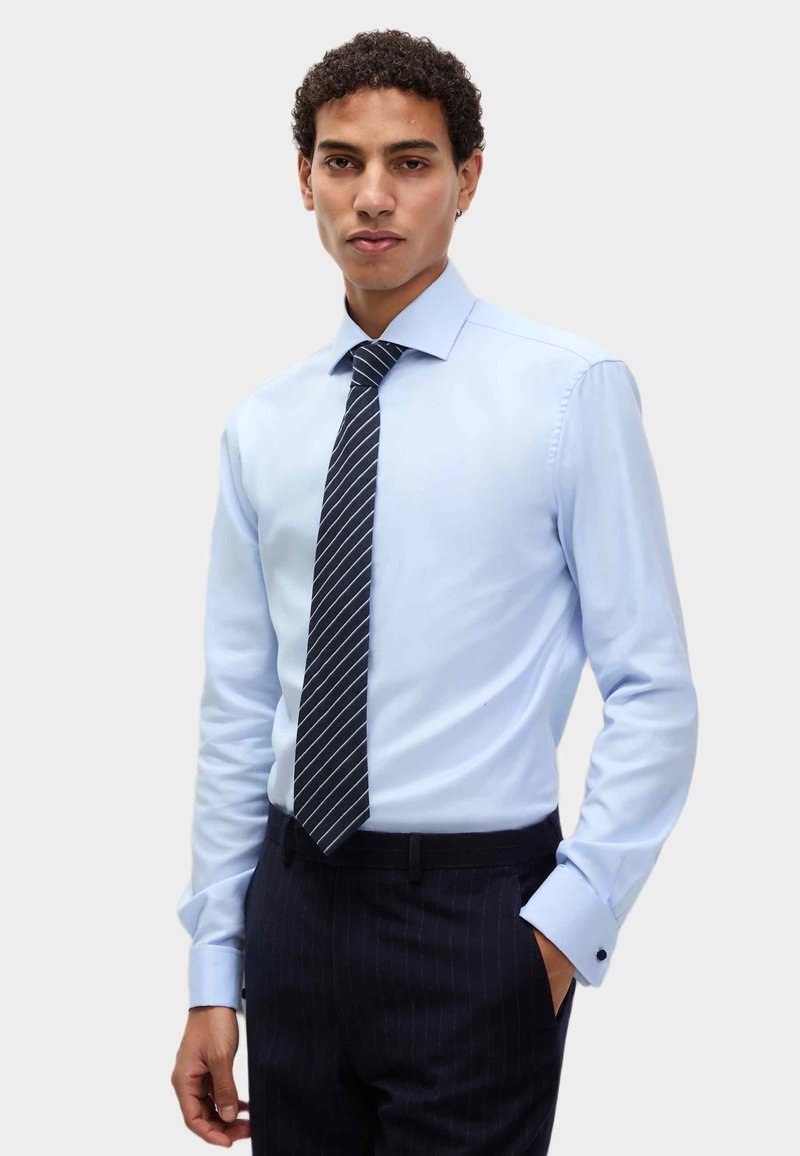 Mężczyzna w jasnoniebieskiej koszuli z czarnym paskiem w paski i ciemnych, prążkowanych spodniach, stojący z jedną ręką w kieszeni.