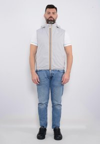 Gilet senza maniche grigio chiaro con colletto alto, cerniera verticale con una striscia gialla e nera, indossato sopra una camicia bianca e jeans blu.
