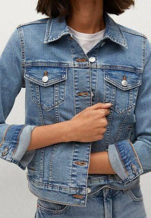 Close-up af en person iført en blå denimjakke med op rullede ærmer over en hvid skjorte, der knapper jakken.