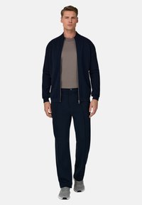 Chaqueta bomber azul marino con cremallera sobre camiseta beige, combinada con pantalones azul marino y zapatillas deportivas grises. Tejido suave y ajuste entallado.