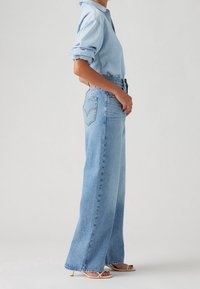 Femme portant une chemise en jean bleu clair aux manches retroussées et un jean bleu taille haute, jambes larges, avec des sandales à talons beiges à bout ouvert.