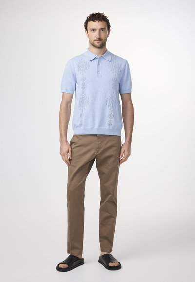 Vaaleansininen lyhythihainen polo-paita, jossa on teksturoituja kuvioita, yhdistettynä beigeihin housuihin ja mustiin liukuviin sandaaleihin. Yksinkertainen napitus kauluksessa.