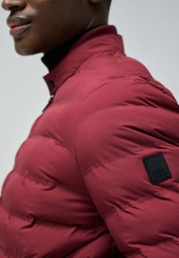 Chaqueta de plumón burdeos con textura acolchada, que presenta un cuello alto y un parche negro con la marca en el brazo izquierdo.