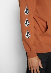 Sweatshirt à capuche orange en coton, avec une poche avant et trois graphismes de logo noirs le long de la manche gauche.