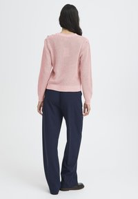 Femme aux longs cheveux noirs portant un pull en tricot rose et un pantalon large navy, se tenant de dos contre un fond uni.